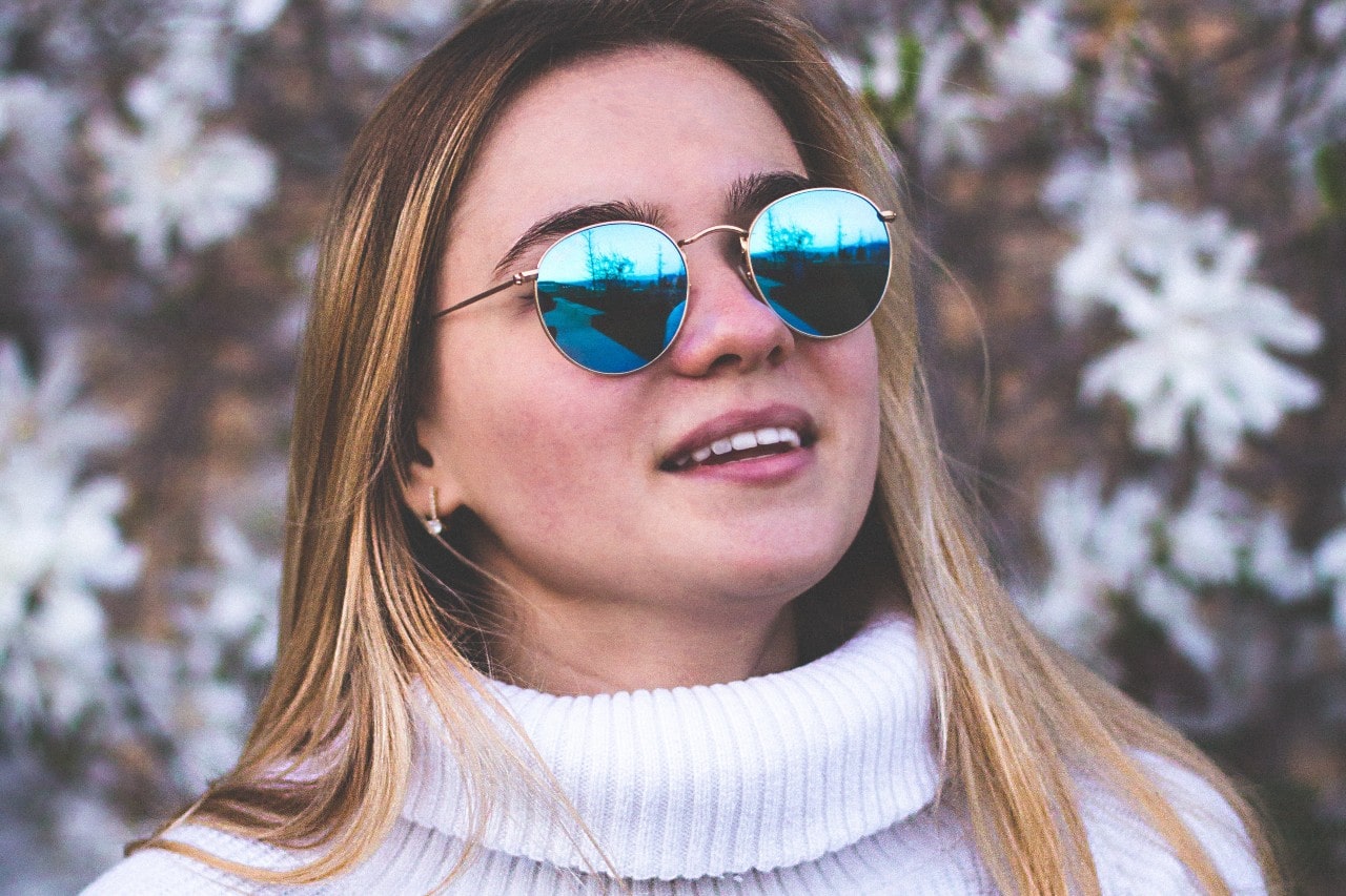 A young woman wearing stud earrings, blue reflective sunglasses and a white sweater, smiling outdoors.