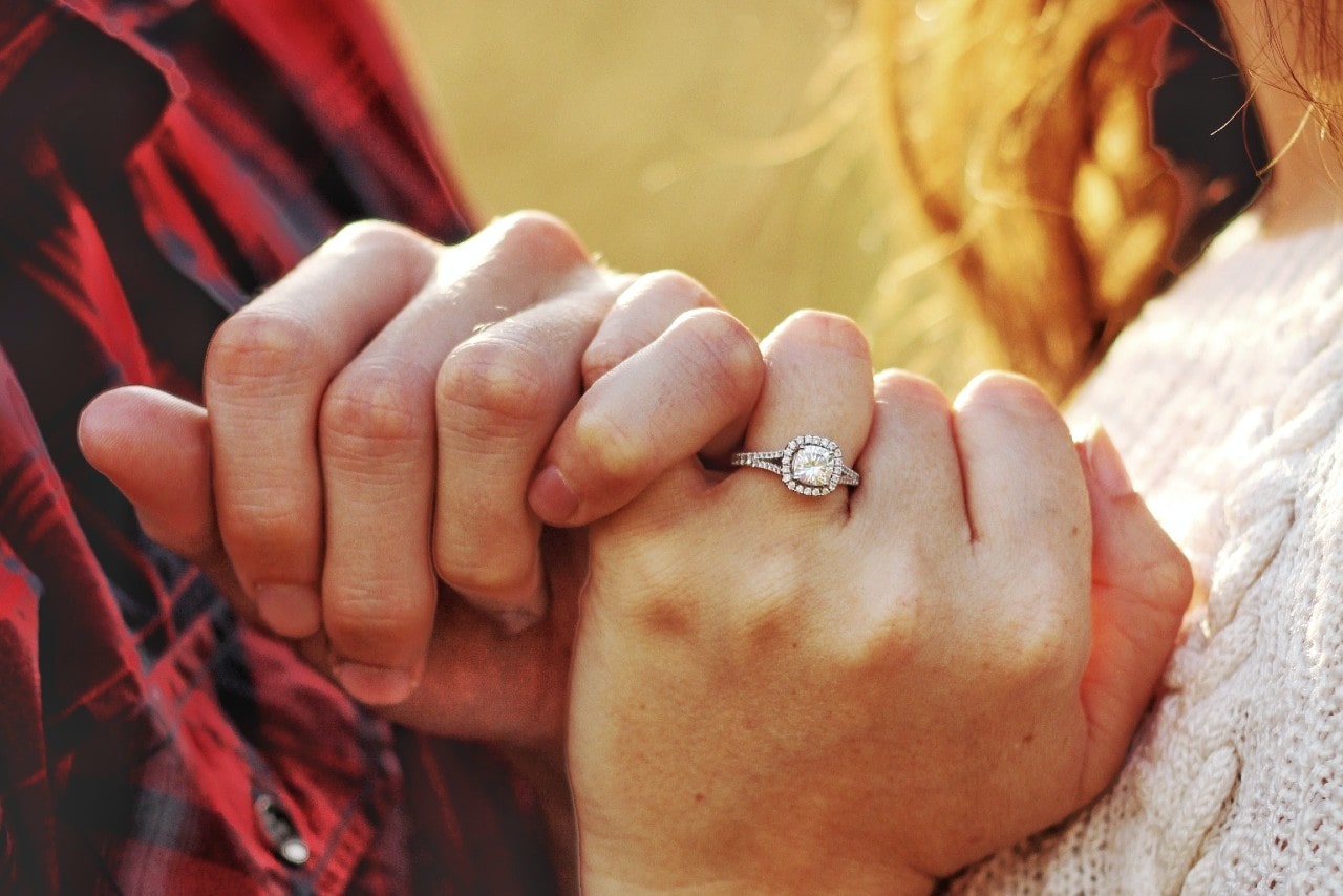 A couple in fall clothing interlocking pinkies, the woman wearing a halo engagement ring