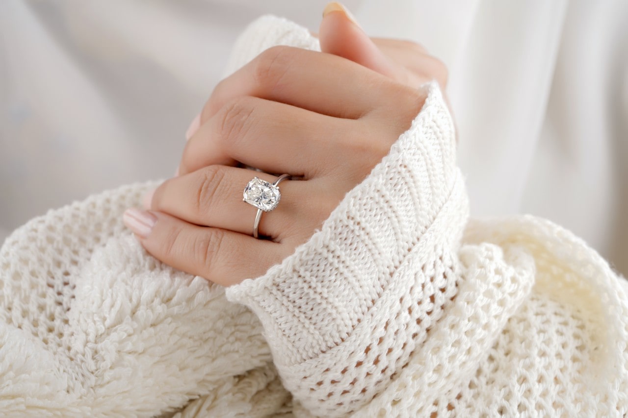 A woman’s hand wrapped in a chunky white sweater sleeve, adorned with a solitaire engagement ring.