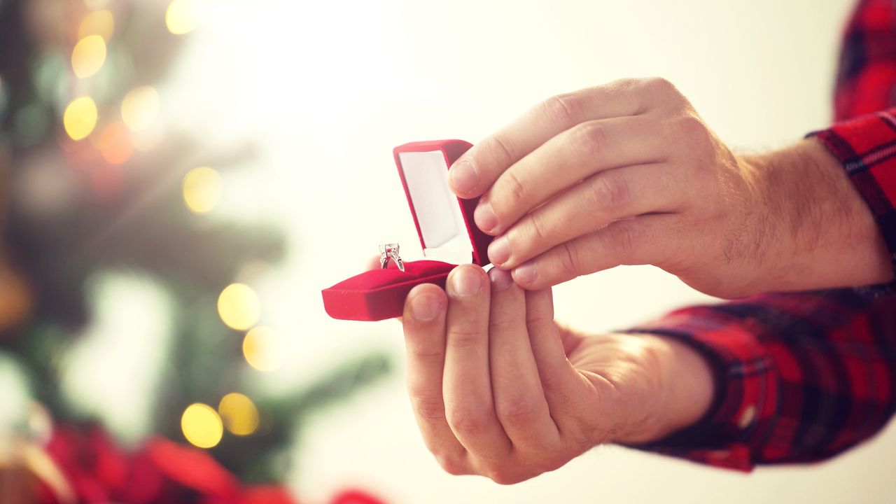 A man in a flannel shirt holding an open engagement ring box with a holiday tree in the background.