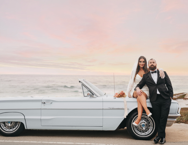 A bride and groom stopped at the beach with the sunset in the background and the bride sitting on the hood of the pale blue Thunderbird convertible.