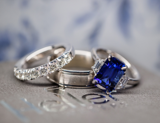 A close-up of a ladies diamond wedding band, an emerald cut sapphire three engagement rings with diamonds flanking the deep blue center stone on top of a brushed metal metal’s wedding band with beveled edge from Heller Jewelers