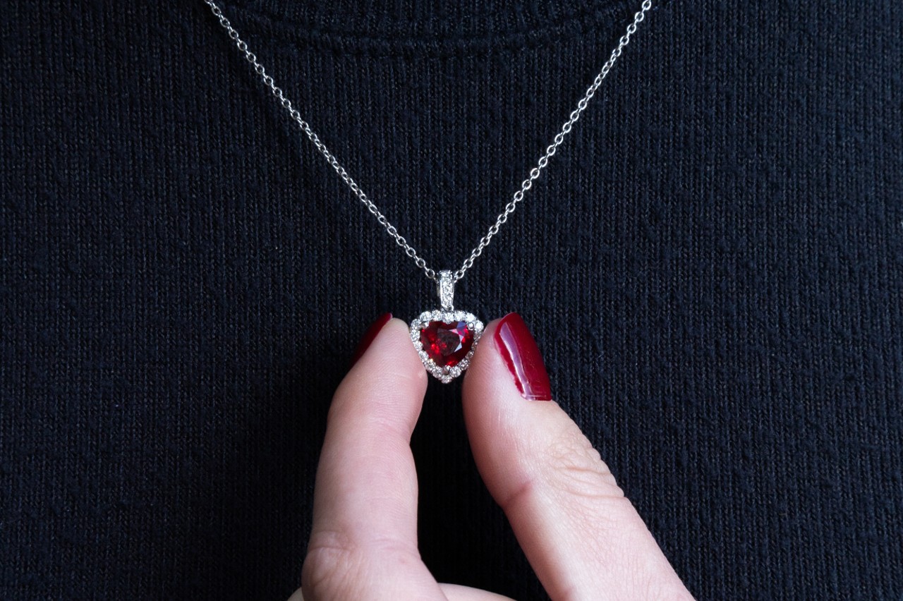 A woman with red nails holding onto the ruby heart pendant with a diamond halo around the heart stone on a white gold chain against her charcoal gray shirt