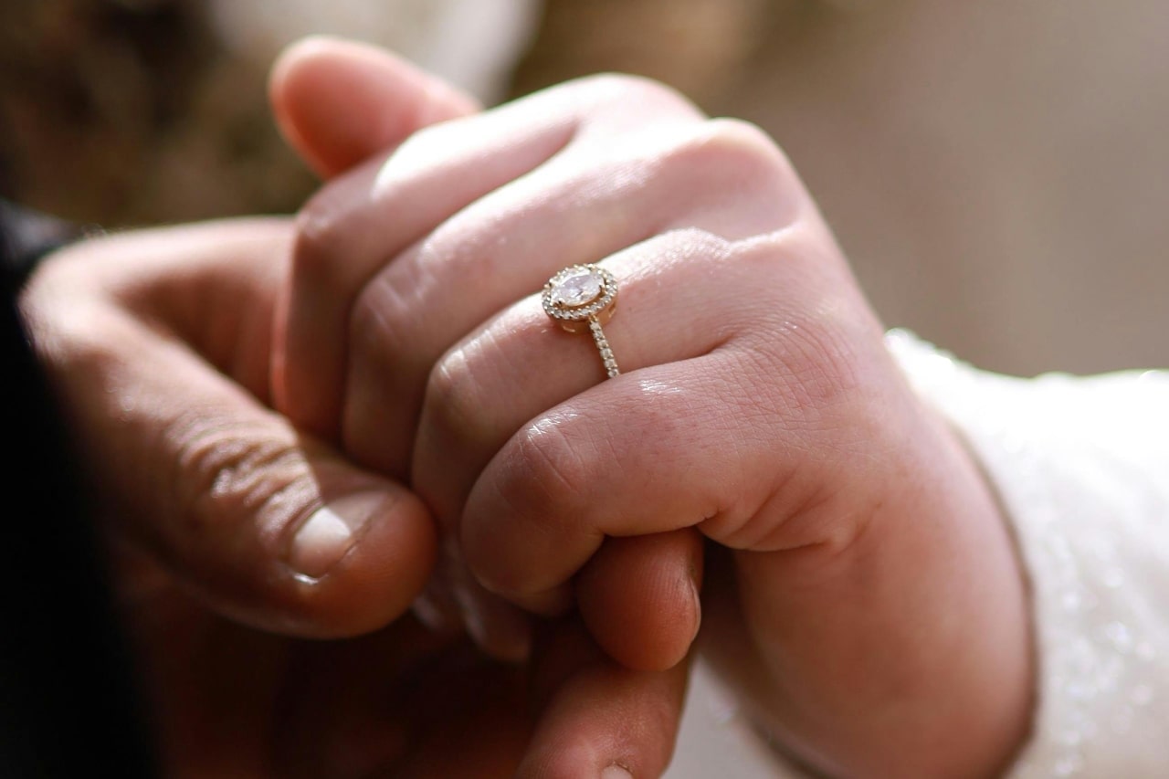 A close-up of a couple holding hands, a halo engagement ring on her finger.