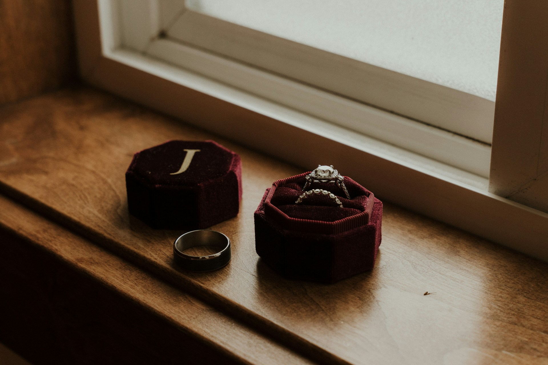 An open ring box on a window sill holding a wedding band and engagement ring, next to a man’s wedding band.