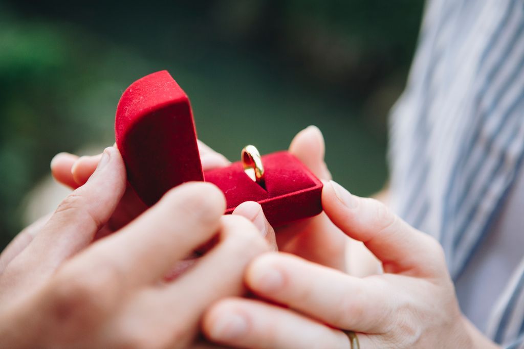 A close-up of a couple holding a red velvet ring box, containing a gold wedding band.