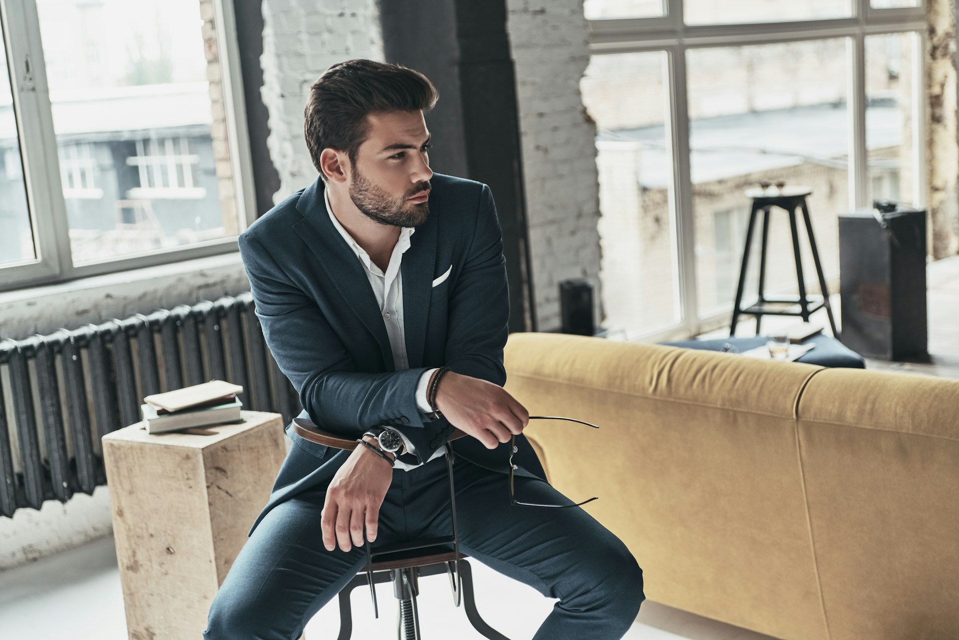 A man sitting on a chair in a chic apartment wearing a suit and luxury watch.